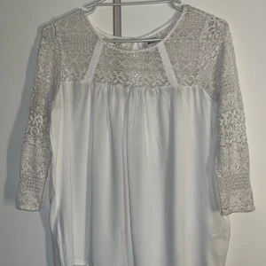 Vit spetsblus från Vero Moda - Supersöt vit blus från Vero Moda med transparent spets upptill och på ärmarna. Blusen har trekvartsärm och rund halsringning, samt en liten knapp i nacken. Perfekt för dig som gillar romantiska detaljer och en luftig känsla.