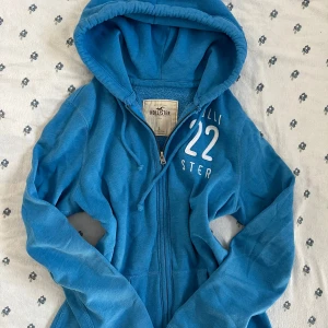 Blå vintage hoodie från Hollister med dragkedja - Säljer en vintage blå hoodie från Hollister med vit brodyr och siffran 22 på bröstet. Tröjan har huva med snörning, dragkedja framtill och två fickor. Tillverkad i mjuk bomullsmix och fodrad med fleece för extra komfort.
