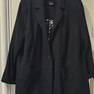 Svart kappa från Lindex Generous - Stilren svart kappa från Lindex Generous med klassisk krage och knäppning framtill. Jackan har stora fickor och rak passform, perfekt för dig som gillar en enkel och tidlös look. Materialet känns mjukt och har ett snyggt fall.