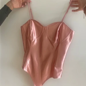 Glansig rosa korsett-topp från H&M - Säljer en snygg glansig rosa korsett-topp från H&M med justerbara tunna axelband och markerade kupor. Toppen har en figurnära passform och är tillverkad i ett syntetmaterial som ger en metallic look. Perfekt för dig som vill sticka ut med en trendig vibe.