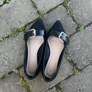 Svarta ballerinaskor med spänne Anna Field - Snygga svarta ballerinaskor från Anna Field med spetsig tå och dekorativt lackat spänne över foten. Skorna har en klassisk och stilren look som passar till många outfits. Tillverkade i syntetmaterial med en platt sula för bekväm känsla hela dagen. 