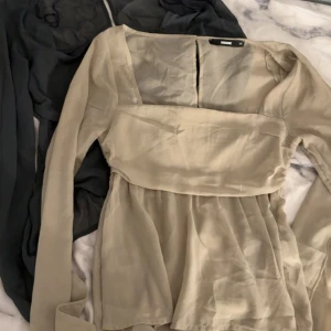 Två transparenta blusar från Bik Bok - Säljer två snygga blusar från Bik Bok i storlek XS. En är ljusbeige och den andra är mörkgrå, båda i transparent polyester med långa ärmar och lätt volang nertill. Den grå har smockad rygg för extra passform. Perfekta att styla med jeans eller kjol.