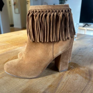Bruna mockaboots med fransar och klack - Snygga boots i brun mocka med bred klack och rund tå. Skorna har coola fransdetaljer runt skaftet som ger en boho-vibe. De är ifrån River island. Aldrig andvända.