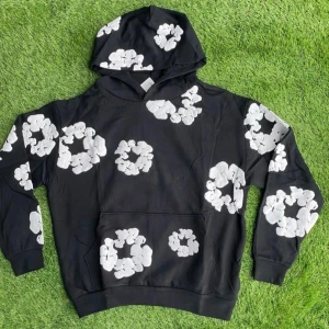 Denim tears hoddie  - Svart hoodie med stora vita blommönster över hela tröjan. Tröjan har huva och en klassisk känguruficka framtill. Perfekt för dig som vill sticka ut med en cool och unik stil.