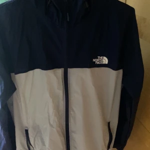 The North Face vindjacka blå/vit - Säljer en snygg tvåfärgad vindjacka från The North Face i marinblått och vitt. Jackan har huva, dragkedja framtill, meshfoder och två fickor med dragkedja. Perfekt för blåsiga dagar och outdoor vibes.