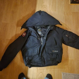  Canada Goose - Svart dunjacka från Canada Goose med huva och klassisk röd-vit-blå patch på ärmen. Jackan har flera fickor, dragkedja och tryckknappar framtill. Perfekt för kalla vinterdagar och har en riktigt schysst streetstil. Bättre pris vid snabb affär såklart 