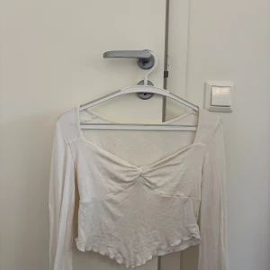 Vit långärmad croppad topp - Säljer en vit långärmad croppad topp med twistad detalj framtill och lätt vågig nederkant. Toppen har en djup, fyrkantig ringning och är tillverkad i ett mjukt, tunt material men är inte genomskinlig