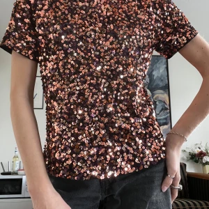 Glittrig paljettopp i koppar och svart - Säljer en kortärmad topp täckt av glittrande paljetter i koppar, orange och svarta toner. Modellen är rak med rund halsringning och har en cool, festlig vibe. Perfekt för dig som vill sticka ut och glänsa! Materialet känns mjukt på insidan och paljetterna ger ett riktigt statement.