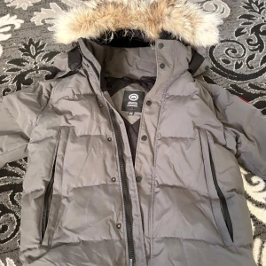 Grå Canada Goose med pälshuva - Säljer en grå dunjacka från Canada Goose med ikonisk patch på ärmen. Jackan har fluffig pälskantad huva, dragkedja och knappar framtill samt två stora fickor med dragkedja. Perfekt för kalla dagar och riktigt snygg streetstil.