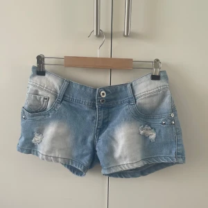 Ljusblå jeansshorts med nitar - Snygga ljusblå jeansshorts med slitna detaljer och coola nitar på fickorna. Klassisk femficksmodell med låg midja och dragkedja. Perfekta för sommaren och ger en avslappnad vibe till din outfit. De är i ett superbra skick! Och har bara används fåtal gånger.