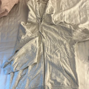 Ljusgrå hoodie med dragkedja XS - Ljusgrå hoodie i storlek XS med dragkedja framtill, huva med snörning och två fickor. Mjuk och skön, perfekt för chill dagar. Klassisk modell med ribbade muddar vid ärmslut och nederkant. Den är oversized