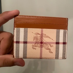 Rutig kortfodral Burb3️⃣rry - Snyggt kortfodral från Burb3️⃣rry i klassiskt rutigt mönster med bruna detaljer. Tillverkat i skinn med tryckt logga och ikonisk ryttare på framsidan. Perfekt för kort och ID, smidig att ha i fickan och ger en lyxig känsla.