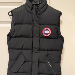 Canada Goose väst - Säljer en svart dunväst från Canada Goose i storlek XS. Västen har klassisk quiltad design, hög krage och två snedställda fickor framtill. Ikonisk Canada Goose-logga på bröstet och tryckknappar längs hela framsidan. Perfekt för lager-på-lager under kyliga dagar. Knappt använd. 