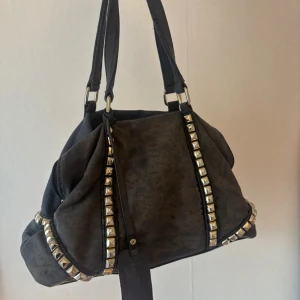 Svart axelväska med nitar från Taska - Cool svart axelväska från Taska med mjuk mockakänsla och silverfärgade nitar längs kanterna. Väskan har dubbla handtag, dragkedja baktill och en snygg tag med logga. Perfekt för dig som gillar en edgy stil och vill ha plats för allt du behöver.