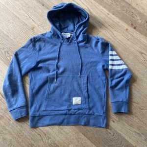 Blå hoodie från Thom Browne med ränder - Snygg blå hoodie från Thom Browne med vita ränder på ena ärmen. Tröjan har huva med snörning, stor magficka och coolt märke framtill. Tillverkad i 100% bomull, perfekt för chill dagar. Passformen är normal och ärmarna är långa.