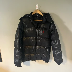 Svart pufferjacka Ralph Lauren M - Svart pufferjacka från Ralph Lauren storlek M. Såklart helt äkta kan skicka bild på kvitto vid intresse. Perfekt jacka till biter som är riktigt varm. Egna negativa är ett litet hål på armen på baksidan av jackan som syns i sista bilden 