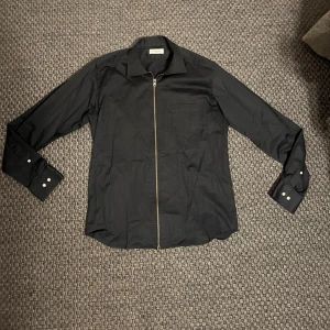 Svart skjorta med dragkedja John Henric - Svart skjorta från John Henric med klassisk krage och hel dragkedja framtill. Skjortan har långa ärmar med manschett och bröstficka. Snygg och stilren design, perfekt för dig som vill ha något lite annorlunda än vanliga knappar.