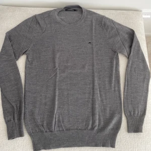 Grå merinoulltröja från J.Lindeberg - Snygg grå tröja från J.Lindeberg i mjuk merinoull. Klassisk rund halsringning, diskret logga på bröstet och ribbade muddar vid ärmslut och nederkant. Perfekt för lager-på-lager och stilren look.