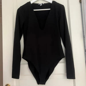 Svart långärmad bodysuit med djup v-ringning - Snygg svart bodysuit med lång ärm och djup v-ringning. Perfekt att styla med jeans eller kjol för en trendig look. Gjord i stretchigt material som sitter tight och bekvämt på kroppen.