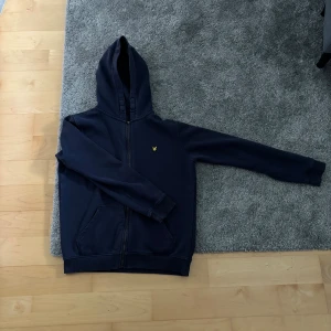 Marinblå hoodie från Lyle & Scott - Säljer en marinblå zip hoodie från Lyle & Scott med dragkedja och huva. Tröjan har en gul broderad logga på bröstet och två fickor framtill. Perfekt för dig som gillar stilren och enkel design. 