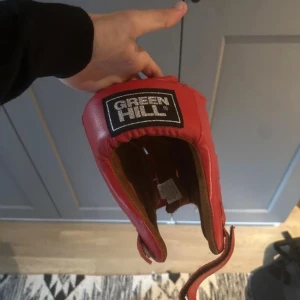 Röd boxningshjälm från Green Hill - Röd boxningshjälm från Green Hill i skinn med vadderad insida och justerbar rem under hakan. Perfekt för dig som tränar kampsport och vill ha extra skydd för huvet. KOM OCH HÄMTA Knivsta (Uppsala)