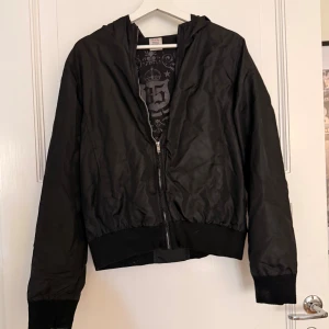 Svart vindjacka  - Svart vindjacka med huva. Jackan har dragkedja framtill, ribbade muddar och en snygg logga på insidan. Lätt och smidig modell som passar perfekt till vardags. Materialet är syntet och jackan har en klassisk, enkel look. Det står ej vilken storlek det är men jag har vanligtvis S så skulle säga så skulle säga att detta passar storlek S. 