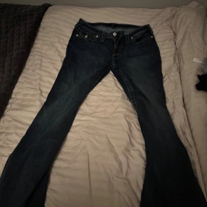 True Religion Joey jeans med bootcut - Säljer ett par mörkblå True Religion Joey jeans med bootcut. Klassiska kontrastsömmar, fem fickor och snygga detaljer på bakfickorna med knappar. Jeansen har låg midja och är tillverkade i bomull med en skön jeanskänsla. Byxorna är helt oanvända 