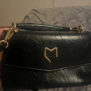 Svart axelväska från Morris - Snygg svart axelväska från Morris, köpt i italien, och ett stilrent M-emblem framtill. Väskan har ett handtag upptill och avtagbar axelrem, perfekt för att bära på olika sätt. Klassisk design som lyfter varje outfit.
