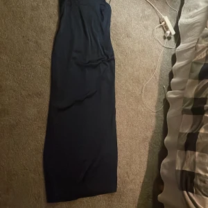 Marinblå långklänning med tunna band - Elegant marinblå långklänning med smala axelband och rak siluett. Klänningen har en minimalistisk design och är tillverkad i ett slätt, lätt glansigt material som ger en lyxig känsla. Perfekt för dig som gillar stilrena och tidlösa plagg. Med en urringning i ryggen 