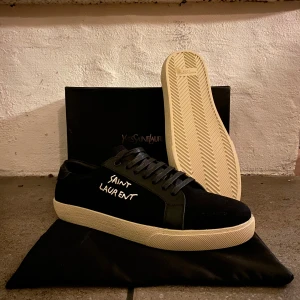  svarta saint Laurent sneakers - Svarta sneakers med vit broderad logga på sidan. Skorna har svart ovandel i canvas och skinn, svarta skosnören och en kontrasterande vit, platt gummisula. Snygg och stilren design som passar till det mesta. Acop