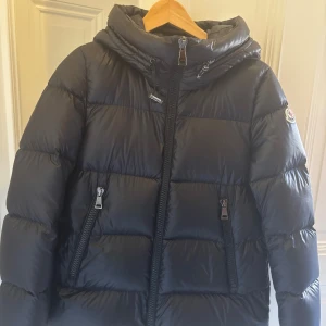 Moncler Serittes Giubbotto - Svart pufferjacka från Moncler med ikonisk logga.  Celebrate Moncler's Serittes down jacket, crafted with glossed leather for a luxurious feel. This regular fit puffer jacket features a short cut, long sleeves, and a practical hood. Designed with a zipper closure and side pockets, it combines padded texture with versatile style.  Kvitto finns.  Nypris 17.000 Storlek TG1 (36-38)