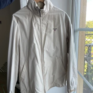 Beige vindjacka från Gant - Snygg beige vindjacka från Gant med dragkedja framtill och två snedställda fickor. Jackan har hög krage och broderad Gant-logga på bröstet. Insidan har tryckt emblem och jackan är tillverkad i ett lätt och smidigt material, perfekt för vår och höst.