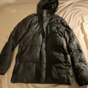 Svart pufferjacka från Calvin Klein Jeans - Säljer en svart pufferjacka från Calvin Klein Jeans med huva och logga på ärmen. Jackan är vadderad och har dragkedja framtill. Perfekt för kalla dagar och har en clean, stilren look som funkar till det mesta.