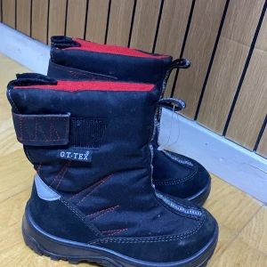 Svarta vinterstövlar från G.T.TEX - Svarta vinterstövlar med röd insida och detaljer, dragkedja framtill och kardborreband på sidan. Grov sula och förstärkta partier i syntetmaterial. Perfekta för kalla dagar och snö.