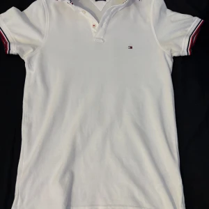 Tommy Hilfiger Piké - Fin och fräsch Tommy Hilfiger Pike. Vit med röda, vita och blåa ränder vid kragen. Bara att komma med funderingar. 