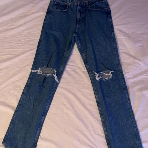 Blå jeans med slitna knän - Säljer ett par klassiska blå jeans med raka ben och snygga slitningar över båda knäna, perfekt för en avslappnad och trendig look. Materialet är robust denim som håller formen.