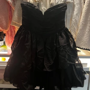 Svart volangklänning från H&M - Supersnygg svart kort klänning från H&M med volangig kjol och axelbandslös design. Klänningen har en markerad midja och flera lager i kjolen som ger extra volym. Perfekt för dig som vill sticka ut med en dramatisk look.