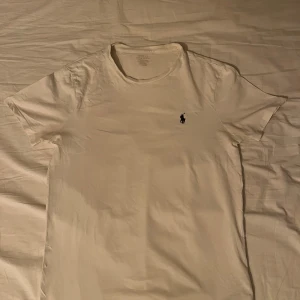 Vit t-shirt från Polo Ralph Lauren - Vit t-shirt från Polo Ralph Lauren med klassisk mörkgrön broderad logga på bröstet. Modellen är enkel med rund hals och korta ärmar, tillverkad i mjuk bomull. Perfekt för en clean och stilren look.