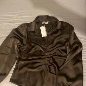 Brun satinblus från Neo Noir - Snygg brun långärmad blus från Neo Noir i glansigt satinmaterial. Skjortan har krage, markerad midja med rynkade detaljer framtill och klassisk knappslå. Perfekt för dig som vill ha en stilren och trendig look. 