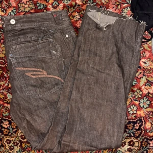 Bruna jeansbyxor med broderad ficka - Säljer ett par bruna jeansbyxor med snygg broderad detalj på bakfickan och råa, fransiga benslut. Klassisk femficksmodell och kontrastsömmar. Perfekta för dig som gillar en avslappnad och unik stil.