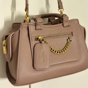 Beige handväska från Vezzano med kedja - Snygg beige handväska från Vezzano med guldfärgade detaljer, kedja och dragkedja framtill. Väskan har både handtag och axelrem, samt en liten tag med logga. Tillverkad i strukturerat syntetmaterial för en modern look & dessutom aldrig använd. 