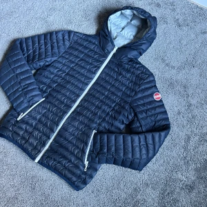 Mörkblå Colmar pufferjacka med huva - Säljer en mörkblå pufferjacka från Colmar med huva och vita dragkedjor. Jackan har quiltad design och Colmar-logga på ärmen. Perfekt för kyliga dagar och riktigt snygg till streetwear-stilen.