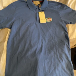 Blå pikétröja från Gucci, storlek L - Snygg blå pikétröja från Gucci med klassisk krage och knäppning framtill. Tröjan har ett broderat Gucci-emblem på bröstet och är tillverkad i mjuk bomull. Perfekt för dig som vill ha en stilren och exklusiv look.