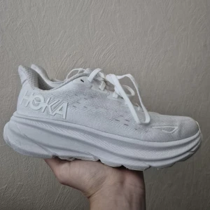 Vita Hoka sneakers med tjock sula - Säljer ett par helvita Hoka sneakers med riktigt chunky sula och ventilerande mesh-ovandel. Skorna har snörning och en sportig vibe, perfekt för dig som gillar komfort och stil. Hoka-loggan syns tydligt på sidan och designen är clean och modern.