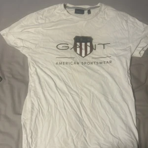 Vit GANT t-shirt med tryck, stl S - Vit t-shirt från GANT med klassiskt tryck framtill och sköldlogga i rött, blått och vitt. Modellen är kortärmad med rund halsringning och tillverkad i mjuk bomull. Perfekt för en clean och sportig look.