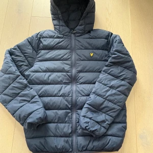 Mörkblå pufferjacka från Lyle & Scott - Säljer en mörkblå pufferjacka från Lyle & Scott Junior med huva och dragkedja. Jackan har det klassiska Lyle & Scott-loggan på bröstet och är quiltad med horisontella sömmar. Perfekt för kyliga dagar och riktigt skön att bära. Fickor på sidorna.