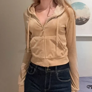 Supersnygg Juicy Couture kofta  - Säljer en beige kofta från Juicy Couture i mjukt tyg med glittrande strassdetaljer på ryggen och logga på bröstet. Tröjan har huva, snörning, dragkedja med J-hänge och långa ärmar. Perfekt för dig som gillar den klassiska Y2K-stilen. Knappt använd! 