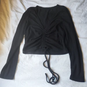 Svart ribbad snörningstopp Gina Tricot - Svart långärmad topp från Gina Tricot med ribbad struktur och croppad passform. Toppen har snygg snörning framtill som kan justeras för att skapa en rynkad effekt. Tillverkad i mjuk polyesterblandning, perfekt för en trendig look.