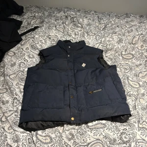 Mörkblå dunväst från Canada Goose - Säljer en mörkblå dunväst från Canada Goose med hög krage och tryckknappar framtill. Västen har två stora fickor, broderad guldfärgad lönnlöv-detalj och klassisk Canada Goose-logga på ena fickan. Perfekt för kyliga dagar när du vill hålla stilen.