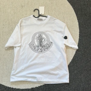 Vit Moncler t-shirt med stor logga - Vit t-shirt från Moncler med stor svart logga tryckt på bröstet och en svart patch på ärmen. Klassisk rund halsringning och avslappnad passform. Tillverkad i mjuk bomull, perfekt för en clean och stilren look.